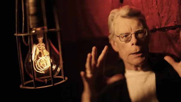 Stephen King, an older white man with pale grey hair and rectangular steel-framed glasses, sits in a red room beside a caged lightbulb.