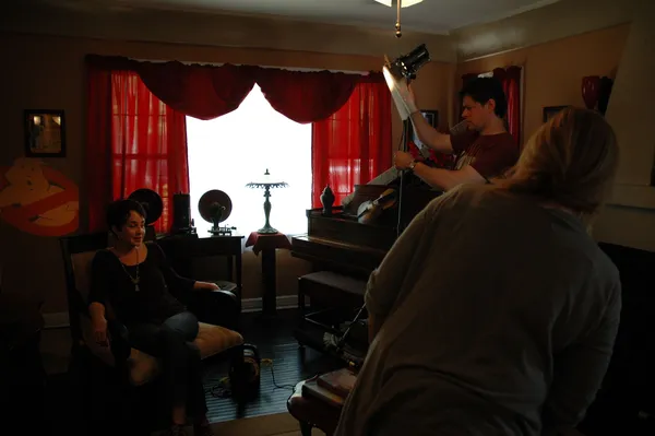 Annie Potts. A slim middle aged white woman in black clothes relaxes in a large chair in front of a red-curtained window as a film crew prepare to start filming her.