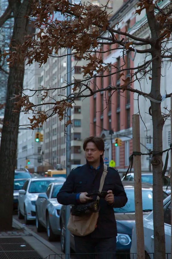 The film director Anthony Bueno. A dark-haired white man wearing a dark blue jacket and carrying a camera as he walks along an urban street lined with autumnal trees.