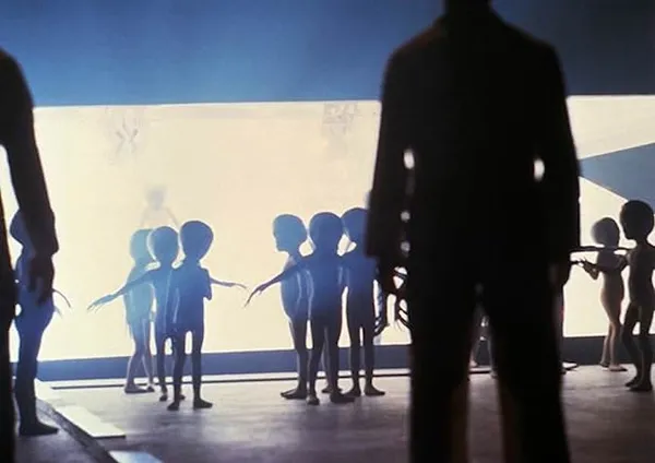 A still from the film Close Encounters Of The Third Kind. A group of small humanoids with long fingers emerges from a spaceship as humans stand nearby.