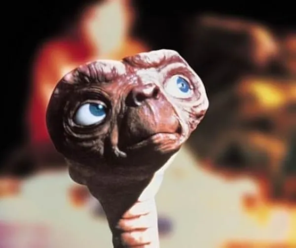 A still from the film E.T. The Extra-Terrestrial. A vaguely humanoid alien with a wide brown head looks up with big eyes.