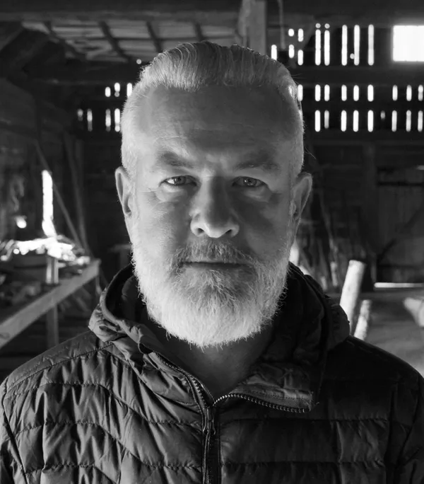 A black and white photograph of the film director Casey Walker. A white man with pale hair and beard, standing in a barn.