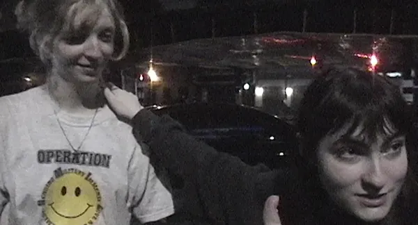 A still from the film Castration Movie Part II. A short, light-skinned, dark haired young poerson looks away while resting a hand on the shoulder of a taller whgite blond wearing a smiley face t-shirt.