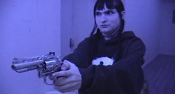 A still from the film Castration Movie Part II. A young person with light-coloured skin, dark hair and black clothing stands in a purple-lit corridor holding a gun.