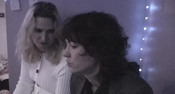 A still from the film Castration Movie Part II. A soart, dark-haired person with light-coloured skin and a taller white person with long blond hair sit side by side.