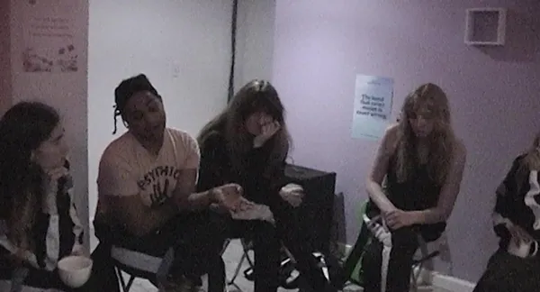 A still from the film Castration Movie Part II. A group of trans women sit in the corner of a windowless room.