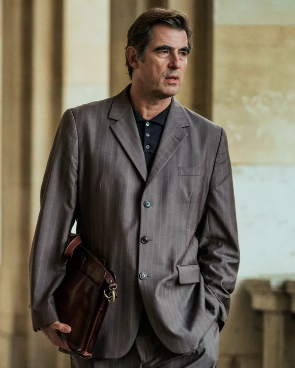 A still from the film The Great Arch. A tall, middle-aged white man with a faintly quizzical expression, wearing a grey suit, clutches a brown leather briefcase under his right arm.