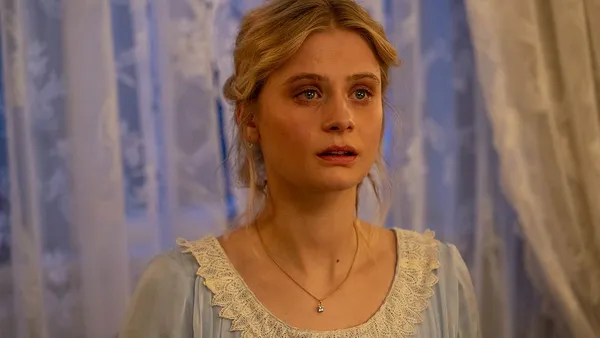 A still from the film The Kiss. A young blond wghite woman stands in front of a net-curtained window, looking worried.