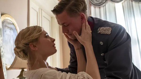 A still from the film The Kiss. A young white man in a military uniform leans down towards a seated young blond white woman, his hand on her neck as she cups his face in her hands. Her left wrist is bruised as if from a restraint.