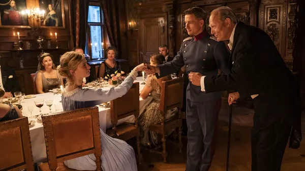 A still from the film The Kiss. At an elegant old-fashioned dinner party, an older white man encourages a white soldier as he takes the hand of a young, blond white woman wearing a long white dress.