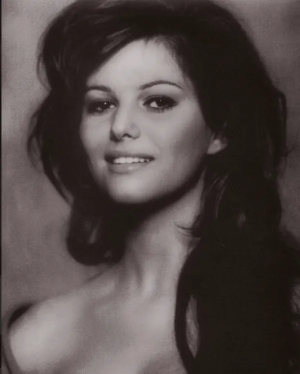 A black and white picture of the actor Claudia Cardinale, taken in her youth. A smiling white woman with long, thick, wavy black hair and dark eyes.