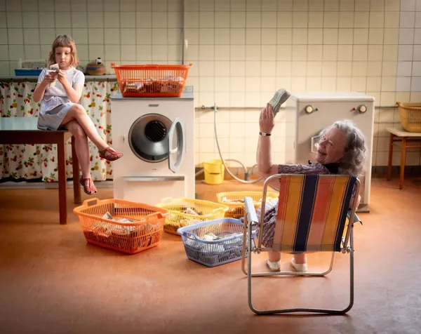A still from the film Two To One. In a laundry room, a young white girl sits on a table counting money, while an old white woman sitting in a folding chair holds up a wad of money and smiles widely.