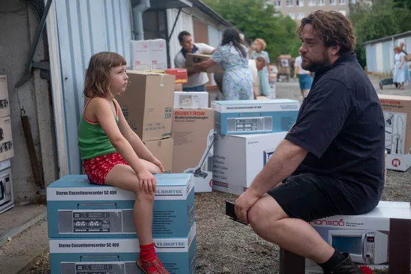 A still from the film Two To One. A young white girl and a white man sit on top of boxes of electronic good, looking at one another, in an outdoor area where other white people are carrying similar boxes around in the background.