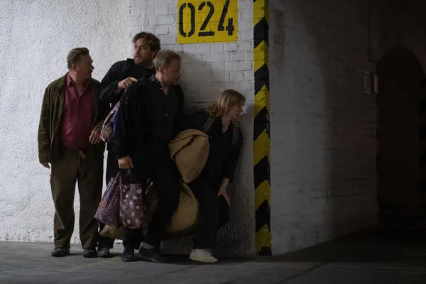 A still from the film Two To One. Four white people carrying large bags sneak along a tunnel, peering cautiously around a corner.