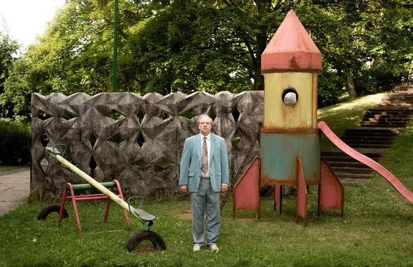 A still from the film Two To One. A white man stands, looking straight ahead, in the middle of a makeshift playground which includes a brightly painted wooden rocket.