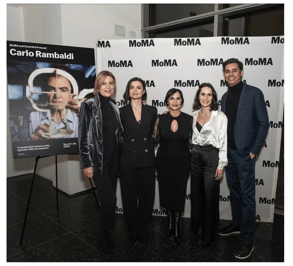Five sharply dressed individuals stand in front of a background on which the word'MOMA' appears repeatedly, beside a poster which says 'Carlo Rambaldi'.
