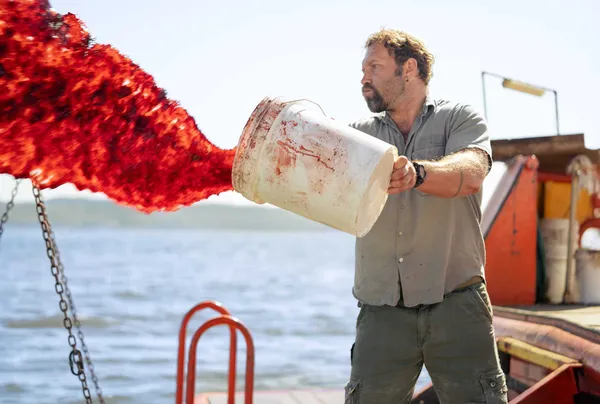 A still from the film Dangerous Animals. A white man throws a bucketload of blood and chum over the side of a boat in a dramatic red spray.