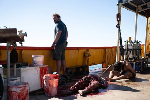 A still from the film Dangerous Animals. A white man stands on the deck of a ship, looking out across the sea. A dead body and a slumped figure in a harness lie at his feet.