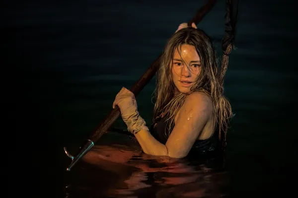 A still from the film Dangerous Animals. A young blond white woman waits in dark water at night. Her left hand is swaddled in cloth and she is holding a spear.