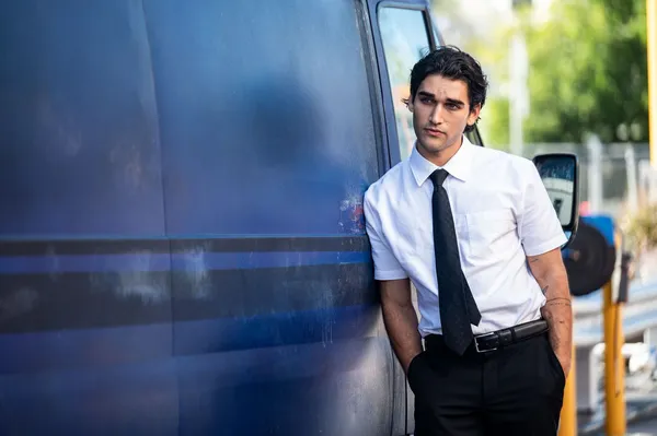 A still from the film Dangerous Animals. A young white man in a shirt and tie stands beside a blue van in a sunny street.