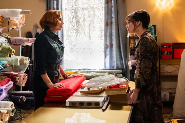 A still from the film Diamanti. Two well-dressed white women exchange stern looks across a wooden counter in a haberdashery, where one is resting her hands atop three bolts of red cloth.