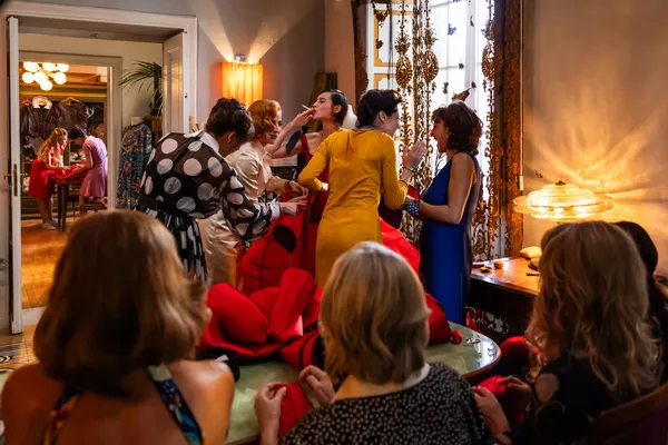 A still from the film Diamanti. A white model tips back her head to smoke a cigarette as four other white woman crowd around her to fit her red gown, with others busily sewing in the foreground.