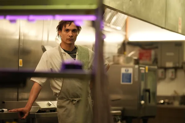 A still from the film Urchin. A young white man stands behind empty shelves in an industrial kitchen.