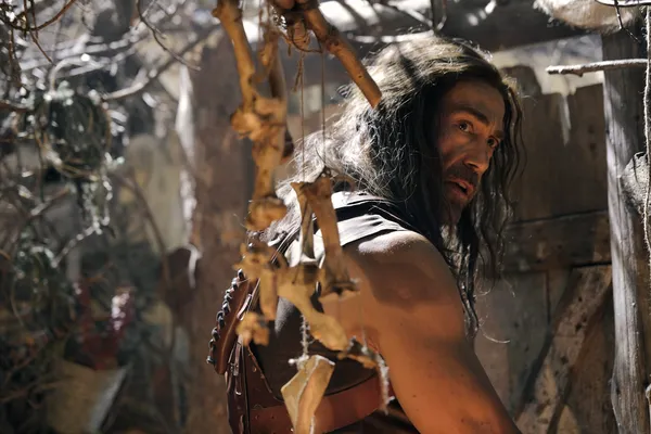 A still from the film Deathstalker. Approaching a wooden door, a long haired white man in pseudo-Medieval warrior clothing notices windchimes made out of bones.