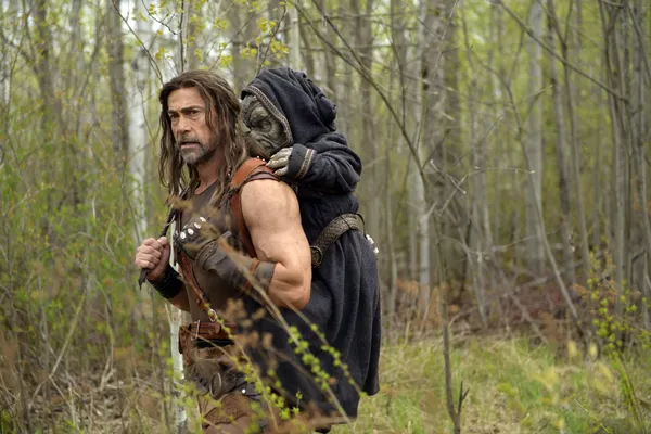 A still from the film Deathstalker. A long-haired white man in pseudo-Medieval warrior's garb walks through woodland carrying a small, black-robed reptilian humanoid on his back.