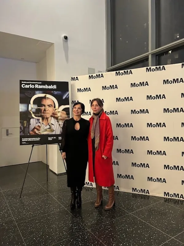 Two middle-aged white women, one in a slender black dress and the other in a log red coat, stand beside a poster advertising a Carlo Rambaldi retrospective.