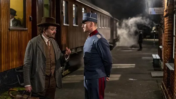 A still from the film The Kiss. A bespectacled middle aged white man in old fashioned clothing, carrying a briefcase and umbrella, talks to a soldier on a train station platform.