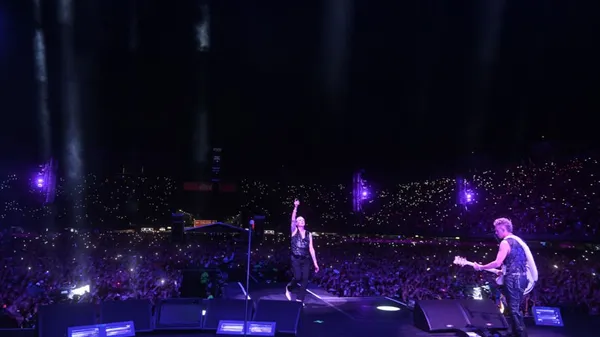 A still from the film Depeche M. The band Depeche Mode, seen from above and behind, swathed in blue and purple light, as they perform onstage to a large audience.