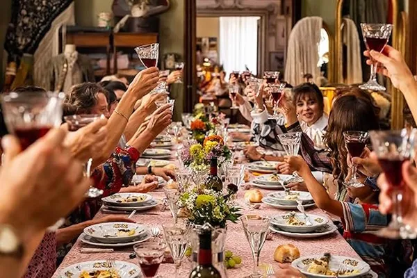 A still from the film Diamonds. A large group of white people lift up wine glasses to make a toast as they sit at either side of a long table laden with food, with an ornate mirror visible through a door at the far end.
