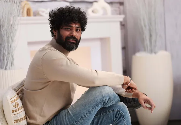 A smiling Maharashtrian man with curly hair and a beard, wearing jeans and a cream-coloured pullover.