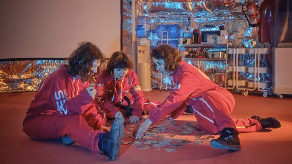 A still from the film Doppelgängers. Three women in red uniforms sit on the floor of a small room with partially metal-paneled walls, assembling a jigsaw.