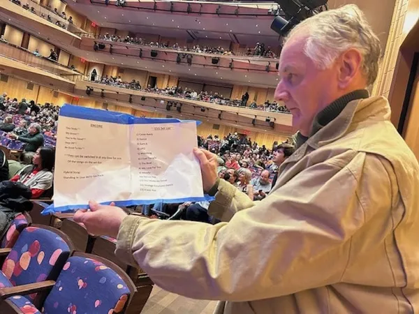 An older white man stands in a busy auditorium, holding up a set list.