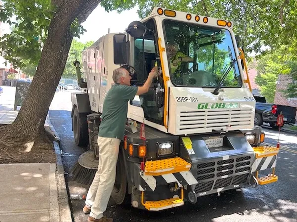An older white man stands by the side of a street cleaning vehicle on a leafy avenue, reaching upb to shake hands with the driver.