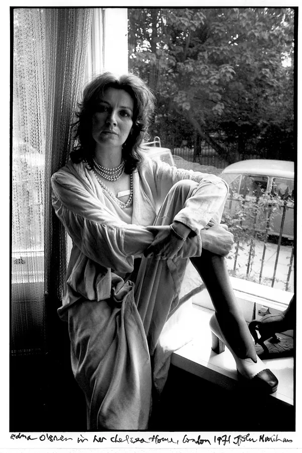 Edna O'Brien, in black and white. A white woman with long, dark, wavy hair, sitting in a tall window with a net curtain hanging at her back and a view of mature trees.