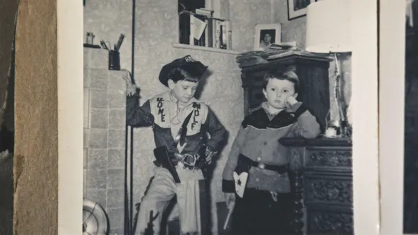 Two white boys in costumes play indoors, beside handsome wooden furniture, in an old black and white photo.