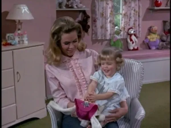 In a pink-walled room, a blonde white woman wearing a pink frilled shirt sits in a pales striped chair holding a smiling blond infant.