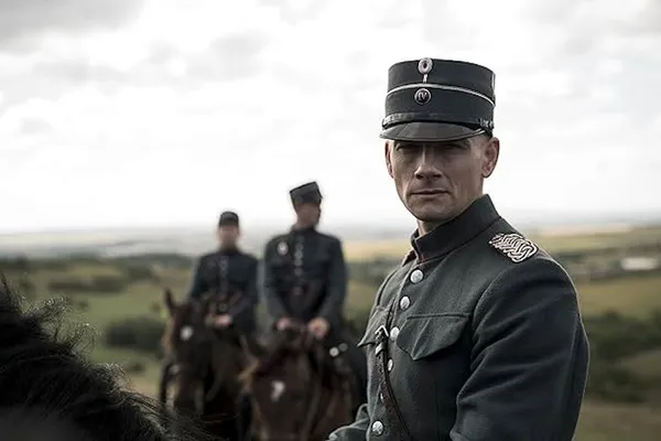 A still from the film The Kiss. A mounted white cavalry officer in an olive drab uniform looks directly at the camera. There are two other mounted white officers behind him, in an open landscape under a moody grey sky.