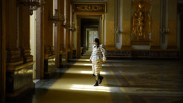 Fabrice Luchini. An older white man in elaborate clothing wanders through a spacious, gilded hall.