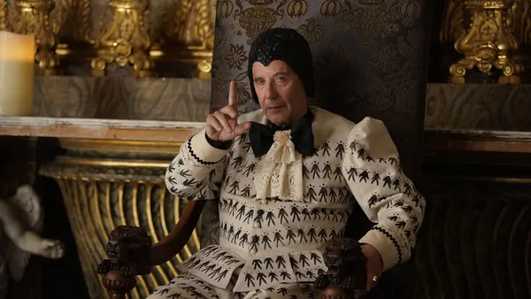 Fabrice Luchini as Belzébuth in The Empire. A white man wearing unusual clothing sits in an elaborate, gilded room.