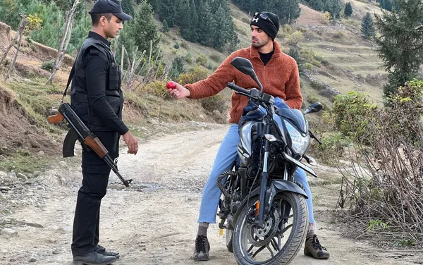 A still from the film We Are Faheem & Karun. On a country road, a young Kashmiri man on a motorbike offers a red apple to a young Kerala man wearing a security guard uniform.