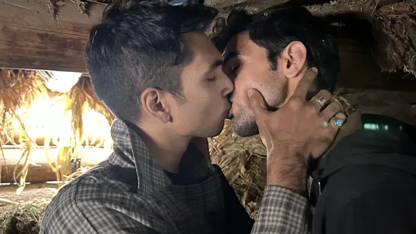 A still from the film We Are Faheem & Karun. A young Kashmiri man and a young Kerala man kiss passionately in a small wooden enclosure in a sunny garden.