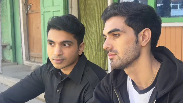 A still from the film We Are Faheem & Karun. A young Kerala man and a young Kashmiri man, both with dreamy expressions, sit side by side on a kerb outside a building with green walls.