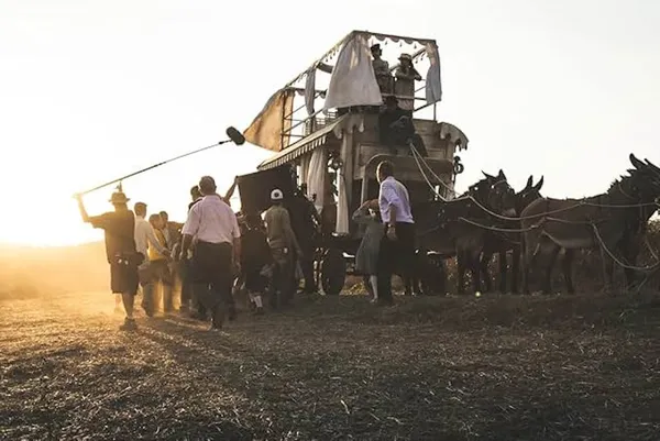 A still from the film The Time It Takes. In a bare field, in evening light, a film crew gathers around a two-level carriage being drawn by four donkeys.