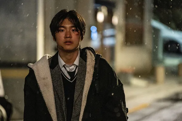 A still from the film Fragment. A young Korean man stands in the street on a snowy night, his face covered in cuts and bruises.