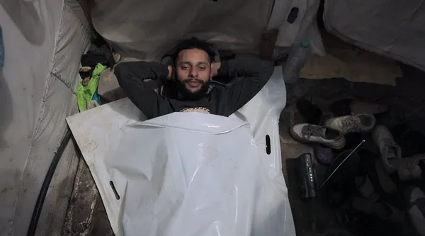 A still from the film From Ground Zero. A Palestinian man lies in a white plastic body bag in a tent, looking directly at the camera.