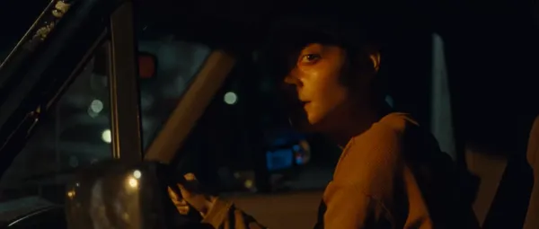 A still from the film Gazer. A young white woman sitting in the driver's seat of a car at night looks to her left as if surreptitiously observing somebody.
